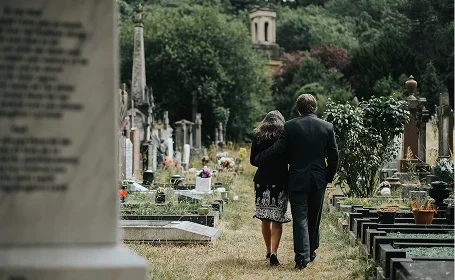 A couple walks closely together through a serene graveyard, surrounded by gravestones and greenery, reflecting a moment of remembrance