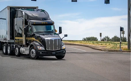 A black semi-truck with a trailer approaches an intersection, under a blue sky with a few clouds and green grass in the background