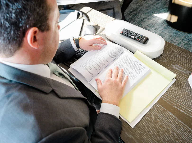 Chris Kinon in a suit, seated at a desk, focused on reading a book or document