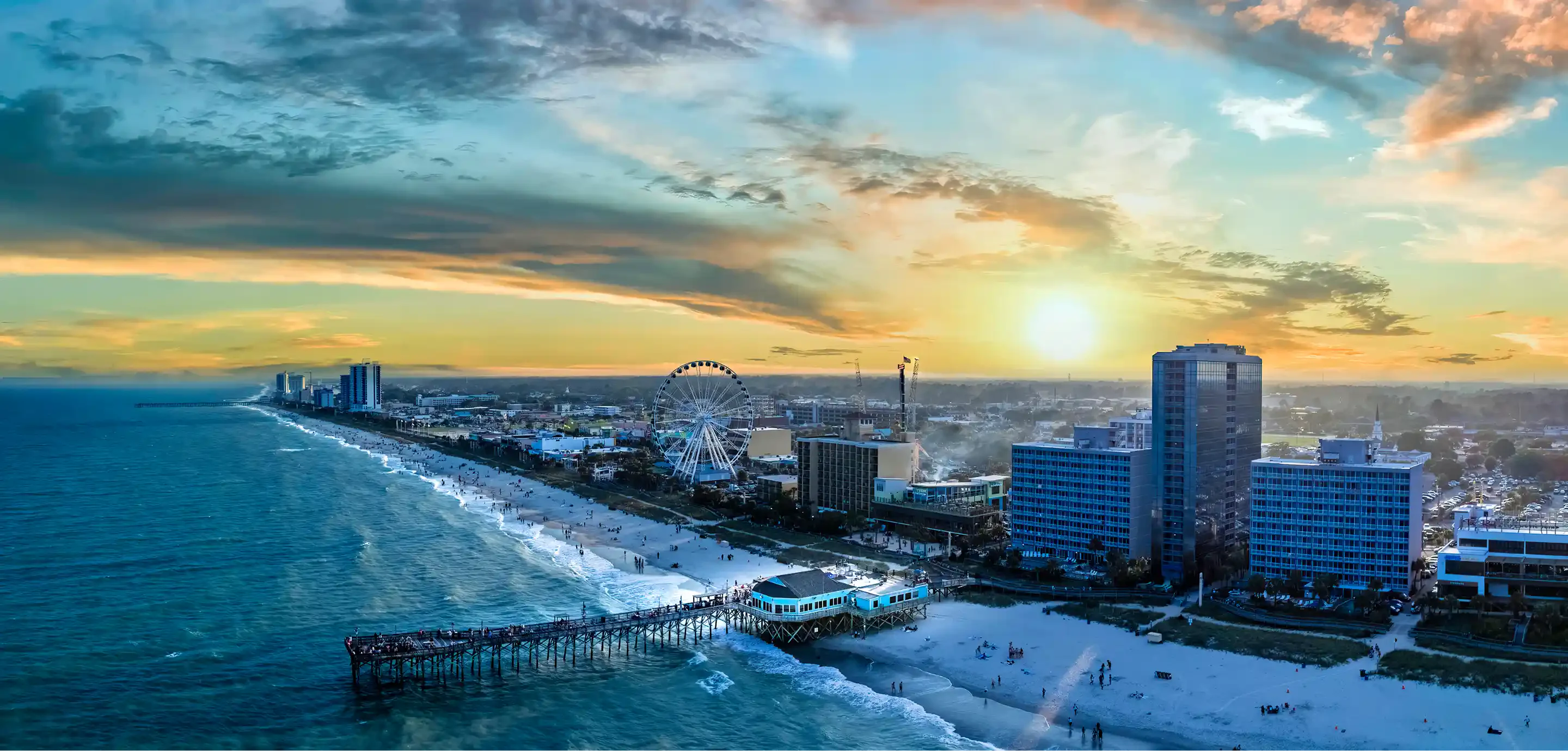 A stunning sunset over a beach city, featuring hotels, a ferris wheel, and a pier along the shoreline with gentle waves lapping at the sand.