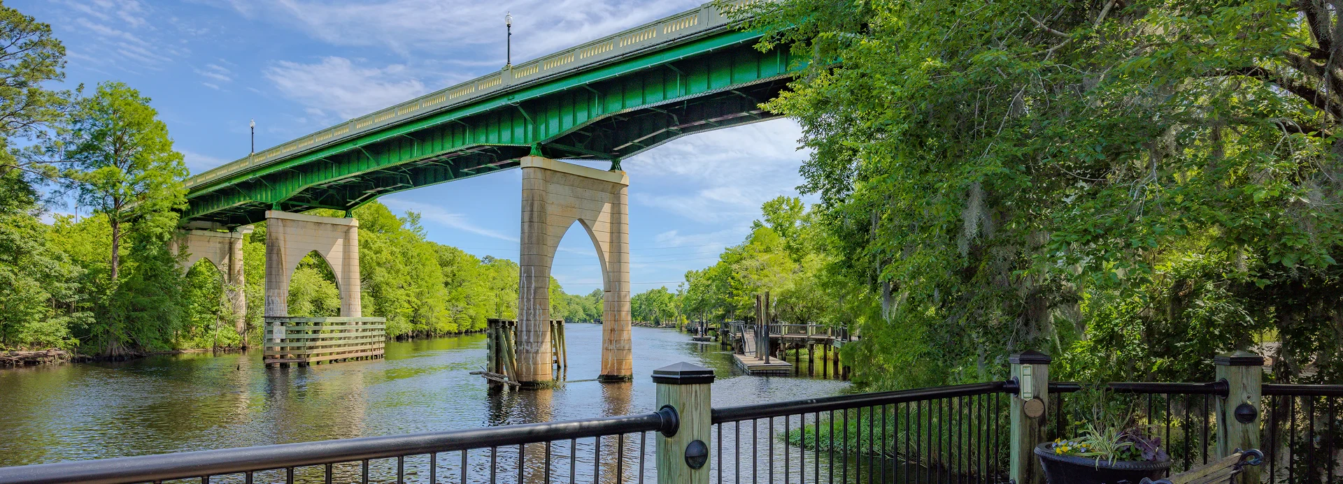 A green bridge spans over a calm river, surrounded by lush green trees and under a bright blue sky, with a wooden walkway in the foreground.